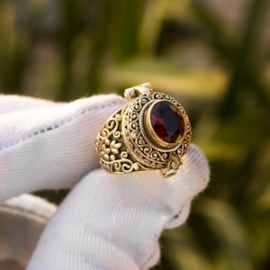 May include: A gold ring with a deep red oval gemstone. The ring has detailed swirling filigree and floral patterns. The ring is held by a gloved hand, highlighting the ornate details and craftsmanship of the jewellery.