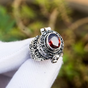 May include: A silver ring with an oval, deep red gemstone. The ring features intricate scrollwork and a hinged top. The gemstone is the focal point, set in a detailed, ornate setting. The ring is shown against a blurred green background.