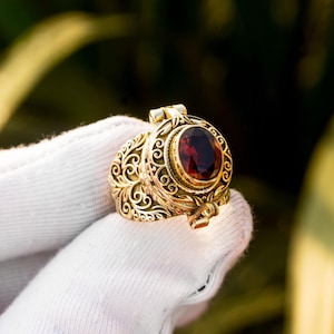 May include: A gold-colored locket ring with an oval, deep red gemstone. The ring features intricate, swirling filigree designs and a hinged top. The gemstone is set in a decorative bezel, adding to the ring's ornate appearance.