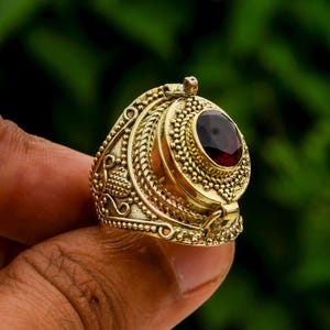 Op de afbeelding: Een gouden ring met een scharnierend deksel en een rode edelsteen. De ring is ingewikkeld versierd met een textuurontwerp.