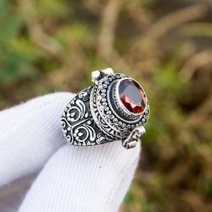 May include: A silver ring with an oval, deep red gemstone. The ring features intricate, ornate detailing around the gemstone and on the band. The ring is held in a white glove, with a blurred green and brown background.