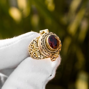 Puede incluir: Un anillo dorado con una piedra preciosa ovalada de color rojo oscuro. El anillo tiene un diseño ornamental y arremolinado alrededor de la piedra preciosa y en la banda. El anillo se sostiene contra un fondo verde borroso.