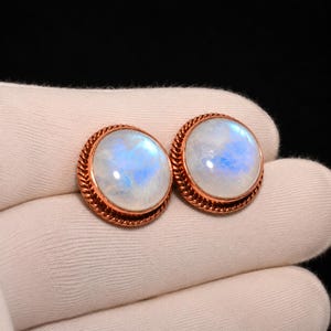 May include: A pair of round stud earrings featuring smooth, translucent moonstones with a blue adularescence. Each stone is set in a polished copper bezel with a decorative twisted rope texture along the outer edge of the circular metal frame.