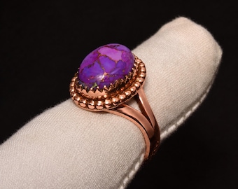 May include: A ring featuring an oval-shaped purple stone with copper-colored veining, set in a polished copper-toned metal band. The setting includes a decorative beaded border surrounding the stone, which is mounted on a split-shank metal band.