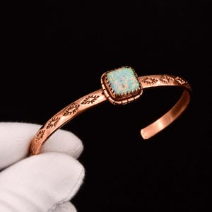 May include: A copper cuff bracelet featuring a square, iridescent white opal stone set in a pronged bezel. The band is decorated with repeating stamped geometric patterns along its length, showcasing a metallic finish and a wide open design.