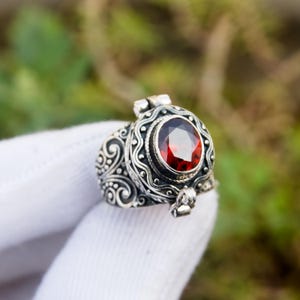 May include: A silver ring with an oval, faceted, red gemstone. The ring has an ornate, detailed design with scrollwork and a hinged compartment. The ring is held up against a blurred green background.