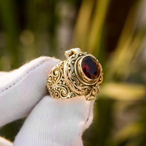 May include: Ornate gold-colored ring featuring a large, oval, deep red gemstone. The ring has intricate scrollwork and dot patterns. The setting is detailed and the ring appears to be vintage or antique in style. The ring is held up by a white gloved hand.