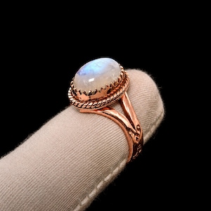 Könnte beinhalten: Ein Kupferring mit einem glatten, ovalen Mondstein-Cabochon, der in einer dekorativen, gedrehten Seilfassung sitzt. Die Ringschiene besteht aus poliertem Kupfer und weist ein geteiltes Design mit feinen Gravurdetails an den Seiten auf.