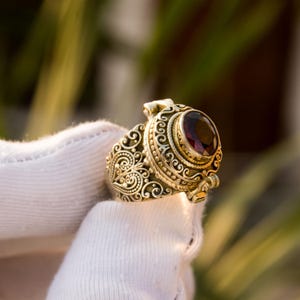 May include: Ornate gold-colored ring featuring a large, oval, deep red gemstone. The ring has intricate scrollwork and filigree detailing around the gemstone and band. The ring is held up by a white glove.