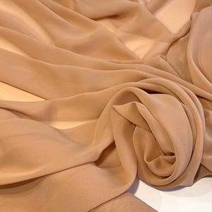 May include: A length of beige fabric, draped and folded to create soft curves and shadows. The material appears lightweight and slightly translucent. A rolled edge is visible at the bottom, suggesting it is a roll of fabric.