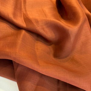 May include: Close-up of a folded, rust-coloured fabric. The material has a subtle sheen and a slightly textured appearance. The fabric is draped, creating soft folds and shadows, highlighting its flowing nature. The overall impression is of a luxurious textile.