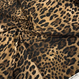 May include: A close-up of leopard print fabric. The fabric has a tan background with dark brown and black spots. The fabric is folded and draped, showing the texture and pattern. The edge of the fabric has the text "100% Co Made in Italy".