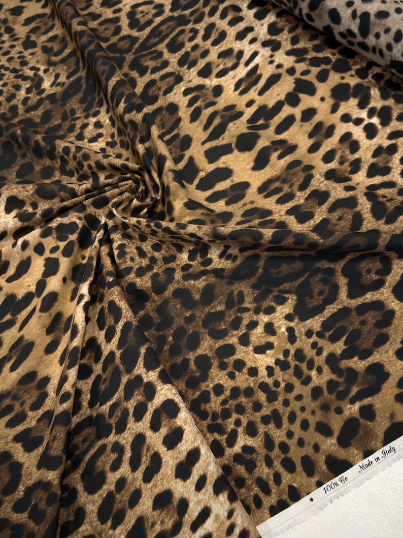 May include: A close-up of leopard print fabric. The fabric has a tan background with dark brown and black spots. The fabric is folded and draped, showing the texture and pattern. The edge of the fabric has the text "100% Co Made in Italy".