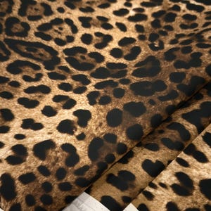 May include: Close-up of a fabric with a leopard print pattern. The fabric features a brown background with black spots of varying sizes. The fabric is folded, revealing a white edge.