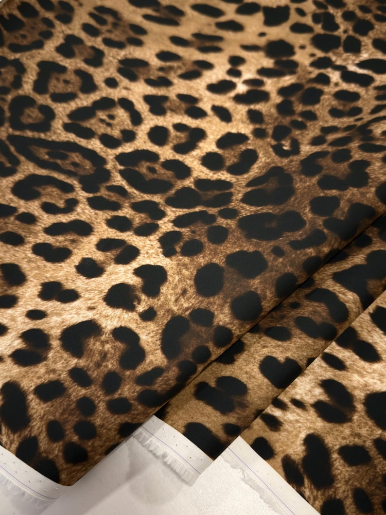 May include: Close-up of a fabric with a leopard print pattern. The fabric features a brown background with black spots of varying sizes. The fabric is folded, revealing a white edge.