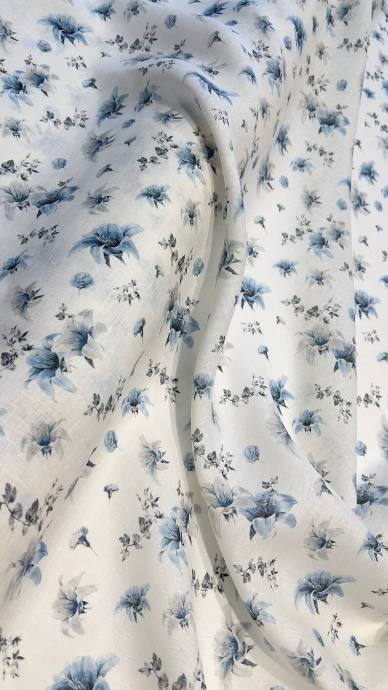 May include: White linen fabric with a blue floral pattern. The fabric is soft and has a subtle sheen. The flowers are small and delicate, and they are scattered randomly across the fabric.