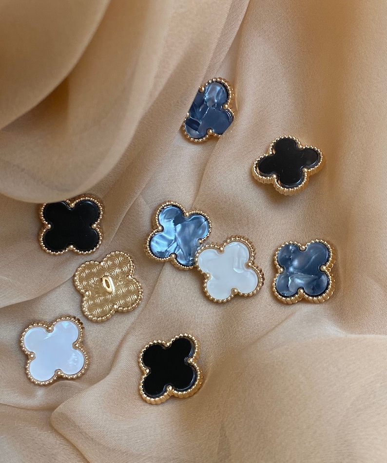 Może przedstawiać: Zestaw ozdobnych guzik&oacute;w w kształcie koniczyny ze złotą obw&oacute;dką. Niekt&oacute;re guziki są czarne, inne białe, a jeszcze inne są marmurkowe szare.