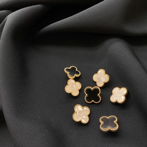 Może przedstawiać: Zbi&oacute;r siedmiu guzik&oacute;w w kształcie koniczyny czteroliściennej. Guziki są wykonane z czarnej i białej żywicy z pozłacanym metalowym obramowaniem.