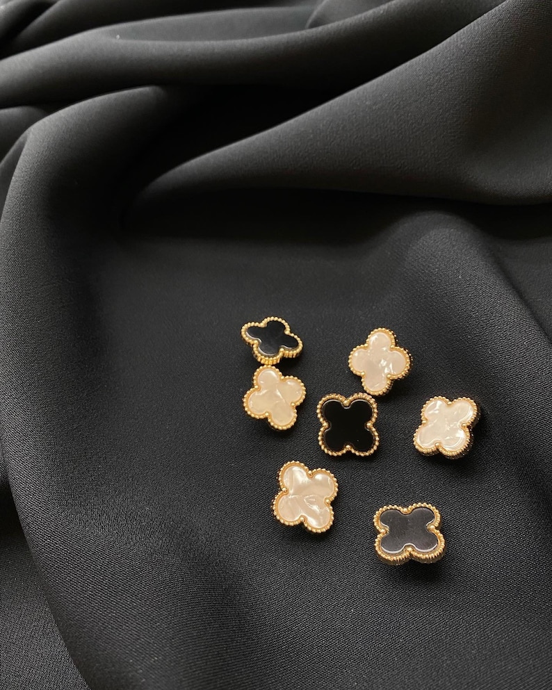 Może przedstawiać: Zbi&oacute;r siedmiu guzik&oacute;w w kształcie koniczyny czteroliściennej. Guziki są wykonane z czarnej i białej żywicy z pozłacanym metalowym obramowaniem.