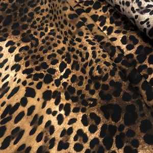 May include: A close-up of a fabric with a leopard print pattern. The fabric has a tan background with black spots and is folded in a way that shows the texture and pattern.