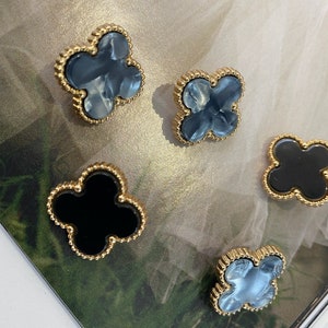 Może przedstawiać: Cztery złote kolczyki w kształcie koniczyny z ozdobną, koralikową obw&oacute;dką. Dwa kolczyki mają wz&oacute;r marmurowy w kolorze niebieskim, a dwa kolczyki mają wz&oacute;r czarny.