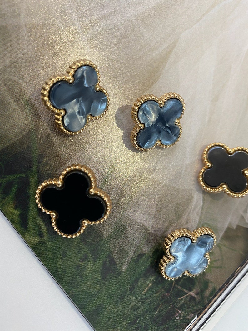 Może przedstawiać: Cztery złote kolczyki w kształcie koniczyny z ozdobną, koralikową obw&oacute;dką. Dwa kolczyki mają wz&oacute;r marmurowy w kolorze niebieskim, a dwa kolczyki mają wz&oacute;r czarny.