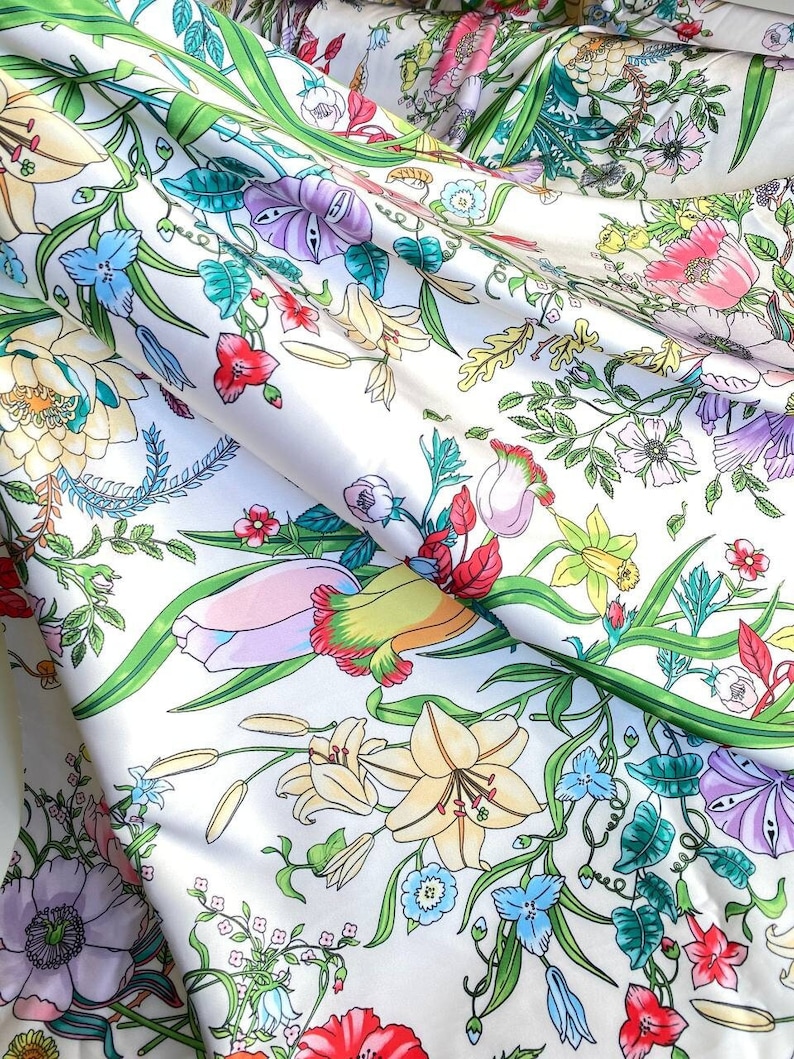 Op de afbeelding: Witte stof met een bloemenprint met verschillende bloemen, waaronder lelies, rozen en madeliefjes. De stof heeft een groene achtergrond en is gemaakt van een zacht, zijdeachtig materiaal.