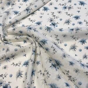 May include: White linen fabric with a blue floral pattern. The fabric is folded and draped, showing the pattern and texture.