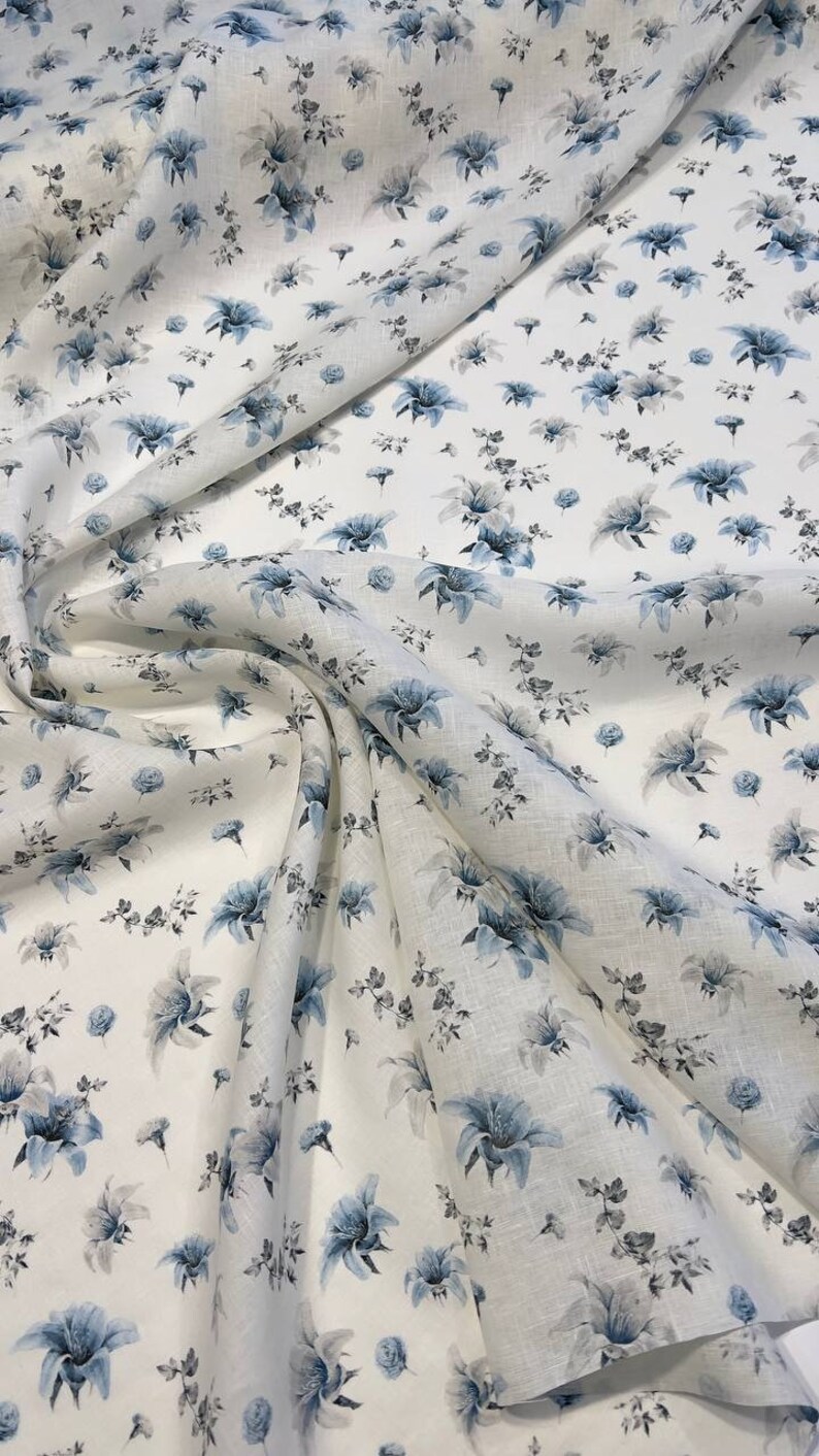 May include: White linen fabric with a blue floral pattern. The fabric is folded and draped, showing the pattern and texture.