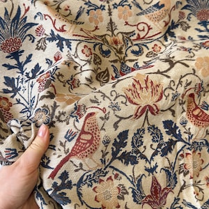 May include: Close-up of a beige fabric with a detailed floral and bird pattern. The design features red, blue, and peach-colored flowers and birds, with dark brown vines and leaves. The fabric is folded, showing the texture and pattern variations.