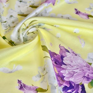 May include: Close-up of a yellow fabric with a floral pattern. The design features large purple and white flowers with green stems and leaves. The fabric has a silky sheen, with folds and shadows adding depth.