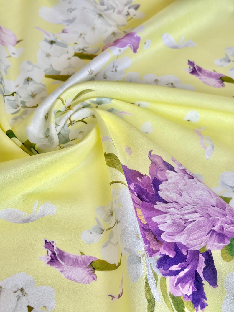 May include: Close-up of a yellow fabric with a floral pattern. The design features large purple and white flowers with green stems and leaves. The fabric has a silky sheen, with folds and shadows adding depth.