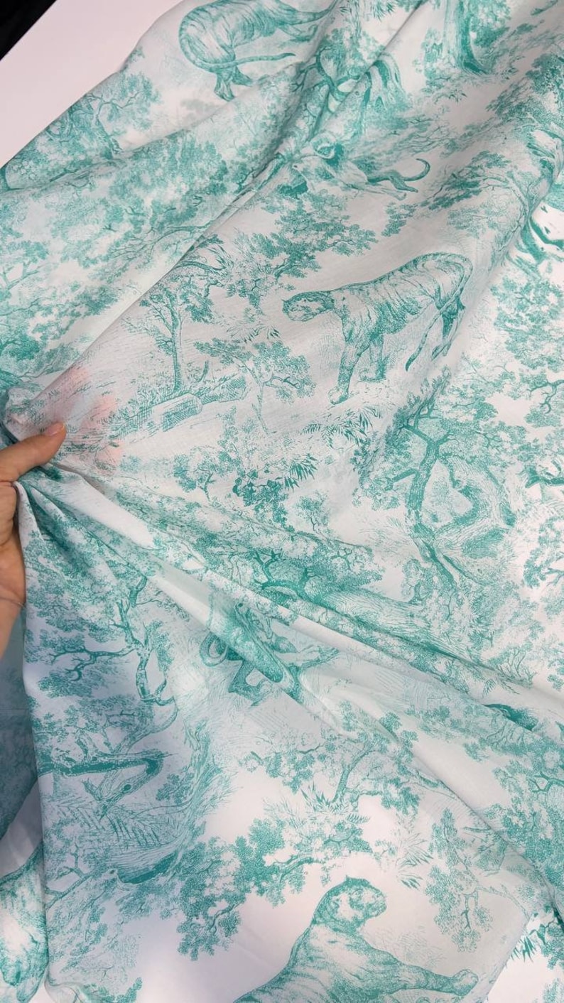 May include: White fabric with a teal green toile pattern featuring tigers, trees, and foliage. The fabric is draped, showing the detailed illustration of a jungle scene. The design is repeated across the material.