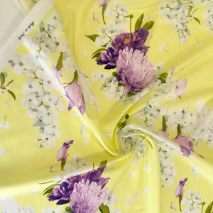 May include: Yellow floral fabric with a repeating pattern of purple and white flowers. The fabric has a satin sheen and is labeled "Made in Italy". The flowers include large, textured blooms and smaller blossoms, set against a bright yellow background.