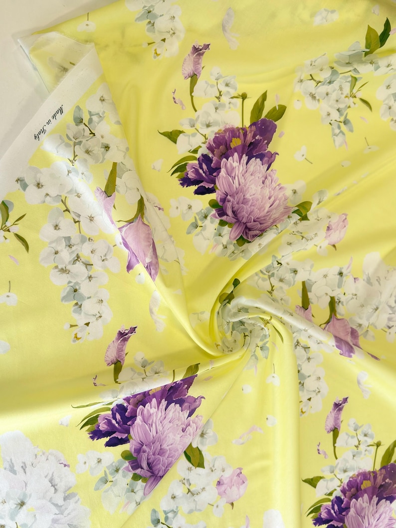 May include: Yellow floral fabric with a repeating pattern of purple and white flowers. The fabric has a satin sheen and is labeled "Made in Italy". The flowers include large, textured blooms and smaller blossoms, set against a bright yellow background.