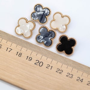 Może przedstawiać: Cztery ozdobne guziki w kształcie koniczyny czteroliściennej. Dwa guziki są białe z perłowym wykończeniem, jeden jest czarny, a jeden szary z marmurowym wzorem. Wszystkie guziki mają złotą metalową obw&oacute;dkę.