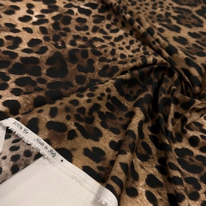 May include: A close-up of a fabric with a leopard print pattern. The fabric is a mix of brown and black spots on a tan background. The edge of the fabric shows a white selvedge with text that reads "100% Co - Made in Italy".