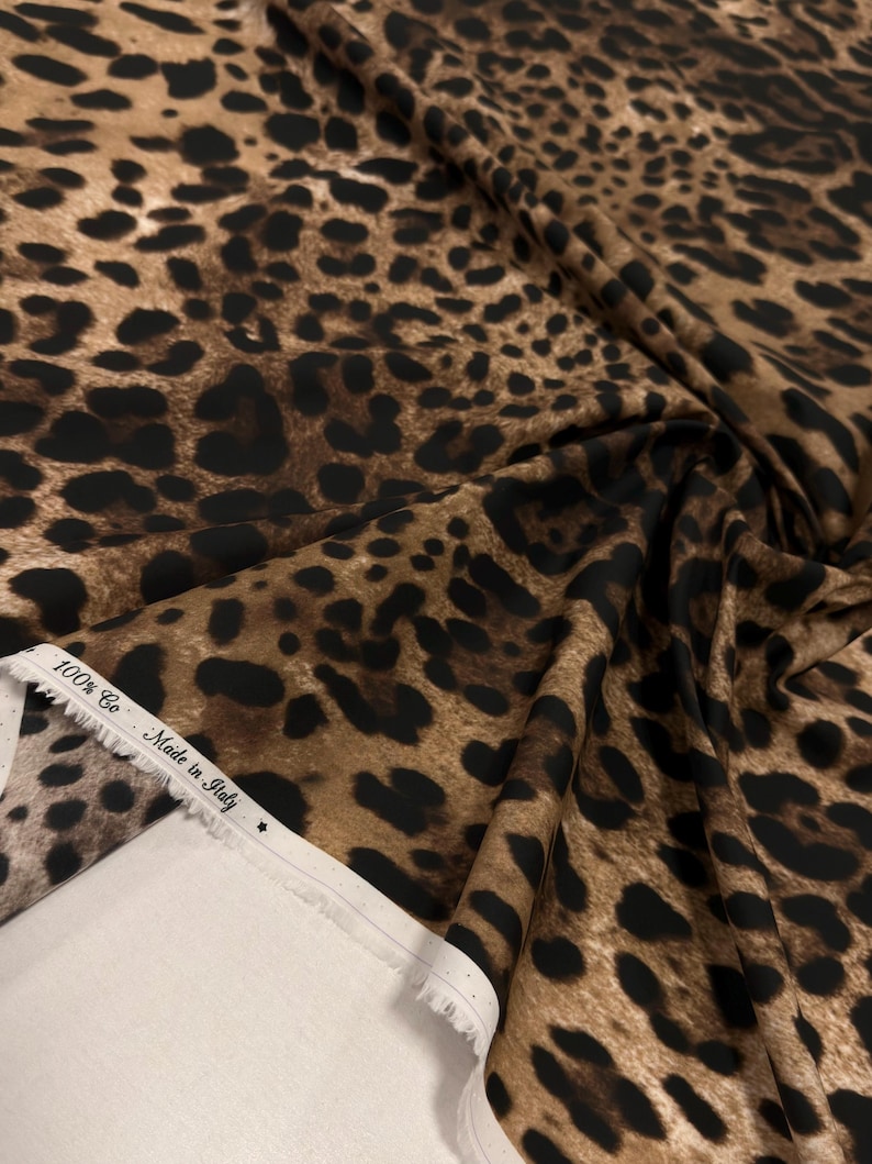 May include: A close-up of a fabric with a leopard print pattern. The fabric is a mix of brown and black spots on a tan background. The edge of the fabric shows a white selvedge with text that reads "100% Co - Made in Italy".