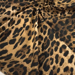 May include: Close-up of a fabric with a leopard print pattern. The fabric features a tan background with dark brown and black spots. The fabric is gathered and folded, creating shadows and texture.