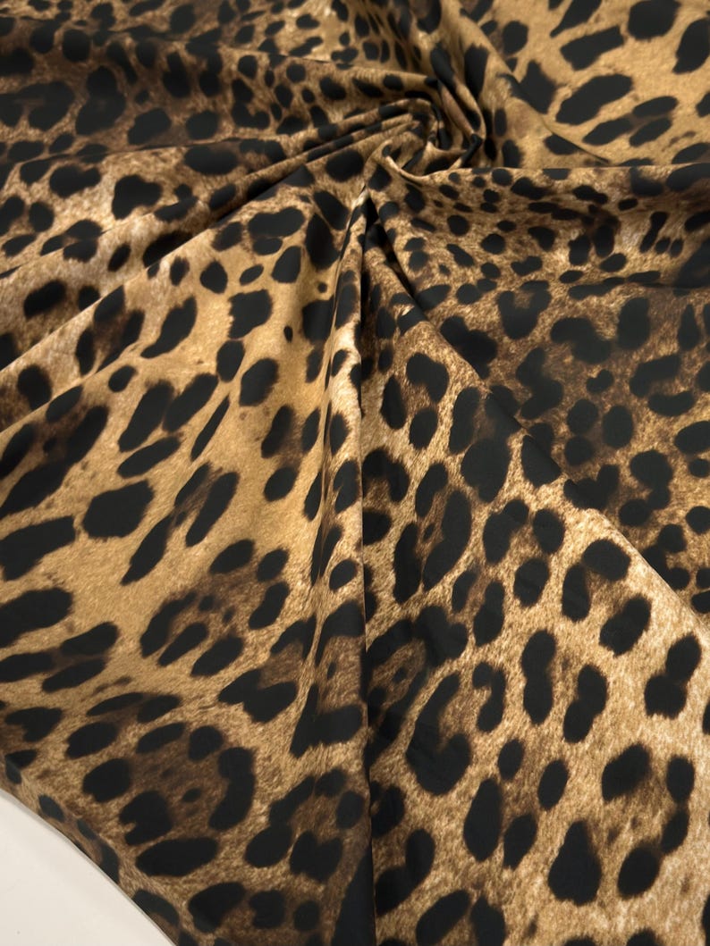 May include: Close-up of a fabric with a leopard print pattern. The fabric features a tan background with dark brown and black spots. The fabric is gathered and folded, creating shadows and texture.