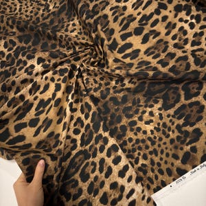 May include: A close-up of a fabric with a leopard print pattern. The fabric features a tan background with dark brown spots. The fabric is folded and draped, showing the texture and design. The edge of the fabric has the text "100% Co Made in Italy".