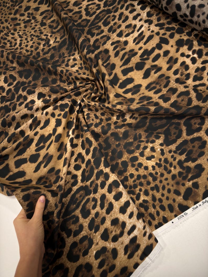 May include: A close-up of a fabric with a leopard print pattern. The fabric features a tan background with dark brown spots. The fabric is folded and draped, showing the texture and design. The edge of the fabric has the text "100% Co Made in Italy".