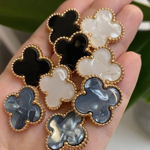 Może przedstawiać: Sześć ozdobnych guzik&oacute;w w kształcie koniczyny z pozłacanym obramowaniem. Guziki są w trzech kolorach: czarnym, białym i szarym.