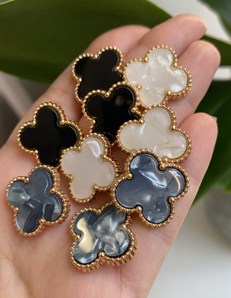 Może przedstawiać: Sześć ozdobnych guzik&oacute;w w kształcie koniczyny z pozłacanym obramowaniem. Guziki są w trzech kolorach: czarnym, białym i szarym.