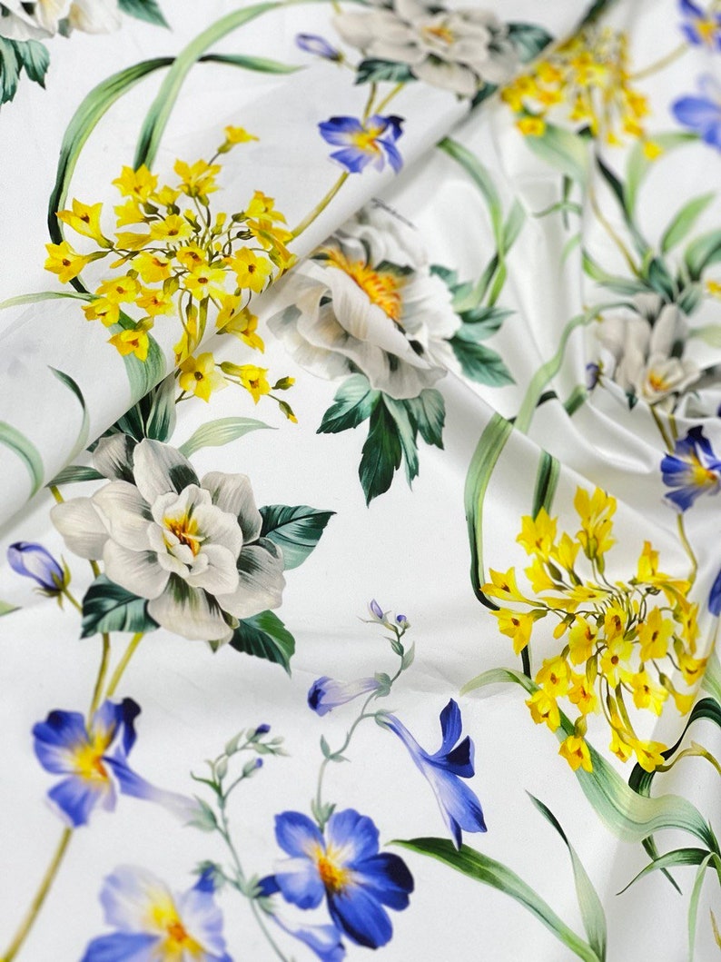May include: White fabric with a floral print featuring yellow, blue, and green flowers. The fabric has a smooth, silky texture.
