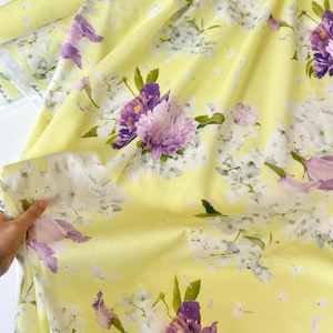 May include: Yellow floral fabric with large purple and light pink flowers, white blossoms, and green leaves. The fabric has a soft, flowing appearance, with a light yellow background. The floral pattern is detailed and vibrant.