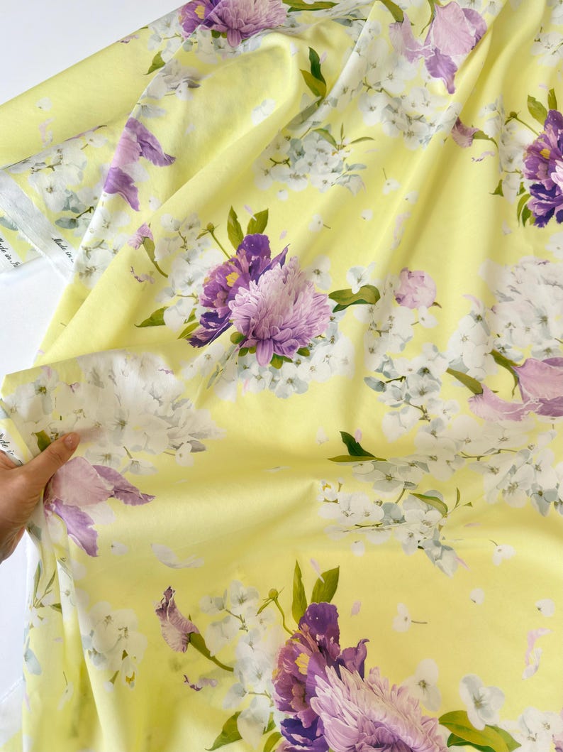 May include: Yellow floral fabric with large purple and light pink flowers, white blossoms, and green leaves. The fabric has a soft, flowing appearance, with a light yellow background. The floral pattern is detailed and vibrant.