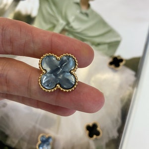 Może przedstawiać: Zbliżenie na niebieski i złoty guzik w kształcie czterolistnej koniczyny. Guzik ma złotą obw&oacute;dkę z małymi koralikami wok&oacute;ł krawędzi. Guzik jest trzymany przez palec.