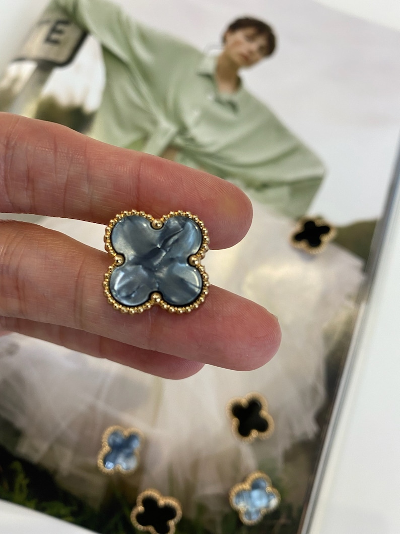 Może przedstawiać: Zbliżenie na niebieski i złoty guzik w kształcie czterolistnej koniczyny. Guzik ma złotą obw&oacute;dkę z małymi koralikami wok&oacute;ł krawędzi. Guzik jest trzymany przez palec.