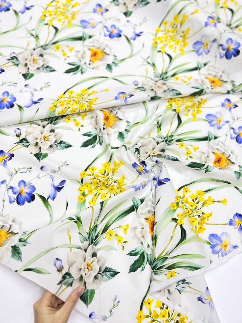 May include: White fabric with a floral print featuring yellow, blue, and white flowers. The fabric has a satin-like sheen.